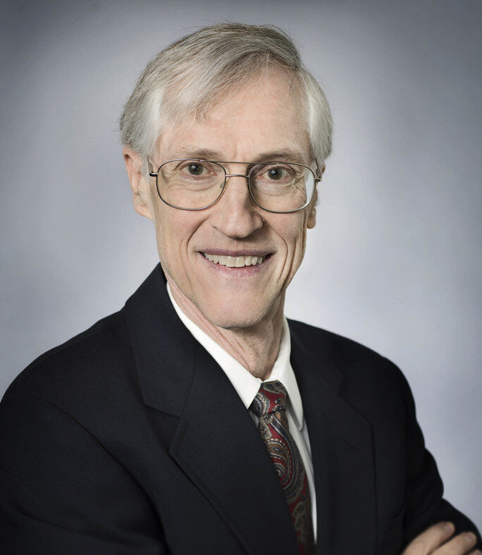 Dr. John Mather smiles warmly at the camera wearing a suit in front of a nondescript background