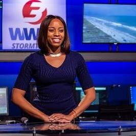 Monique Robinson, a news anchor, stands behind a desk in front of some monitors depicting the news logo