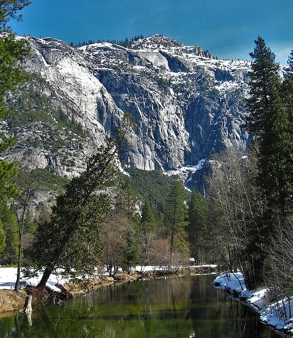A snowy Yosemite mountain behind a lake, also surrounded by snow